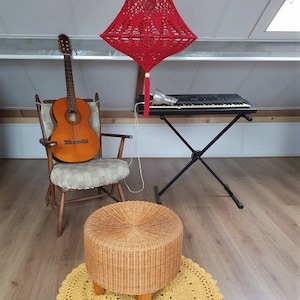 May include: A red crocheted lampshade hangs from the ceiling above a wooden chair with a guitar on it. A black keyboard sits on a stand next to the chair. A wicker stool sits on a yellow crocheted rug.