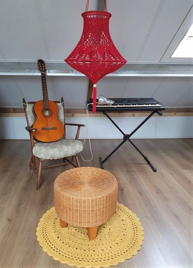 May include: A red crocheted lampshade hangs from the ceiling above a wooden chair with a guitar on it. A black keyboard sits on a stand next to the chair. A wicker stool sits on a yellow crocheted rug.