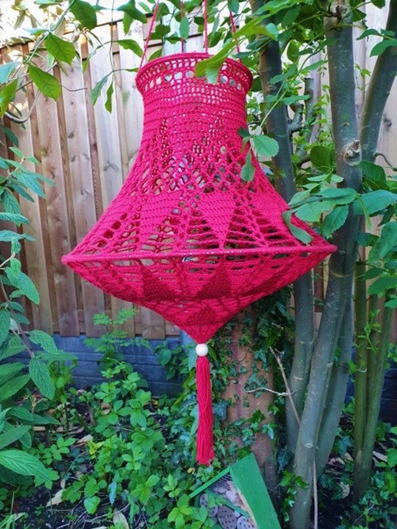 May include: A red crocheted lampshade with a tassel hanging from a tree branch. The lampshade is made of a delicate, openwork design.