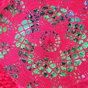 May include: A close-up of a red crocheted doily with a circular pattern. The doily is placed over a green leafy background.