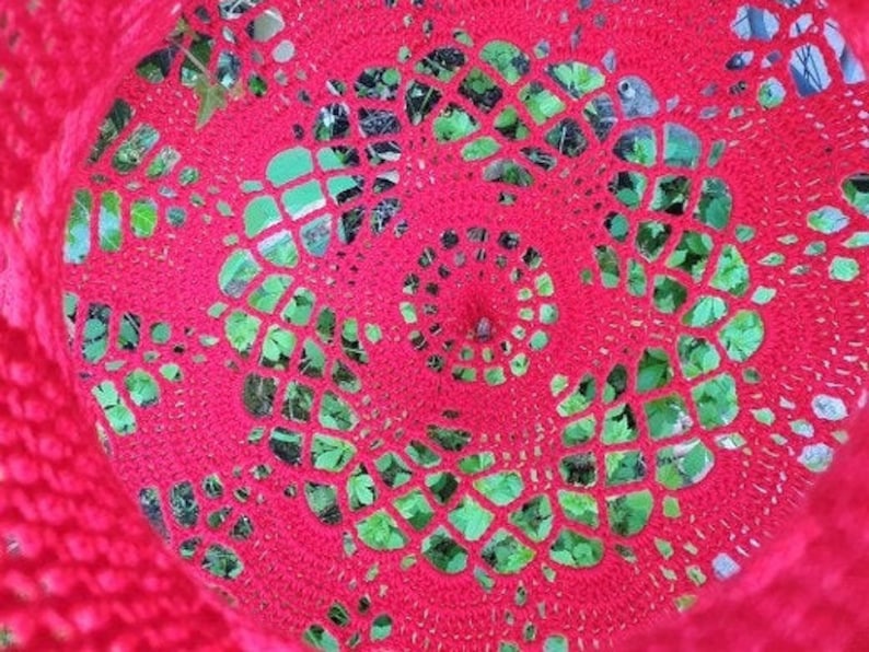 May include: A close-up of a red crocheted doily with a circular pattern. The doily is placed over a green leafy background.