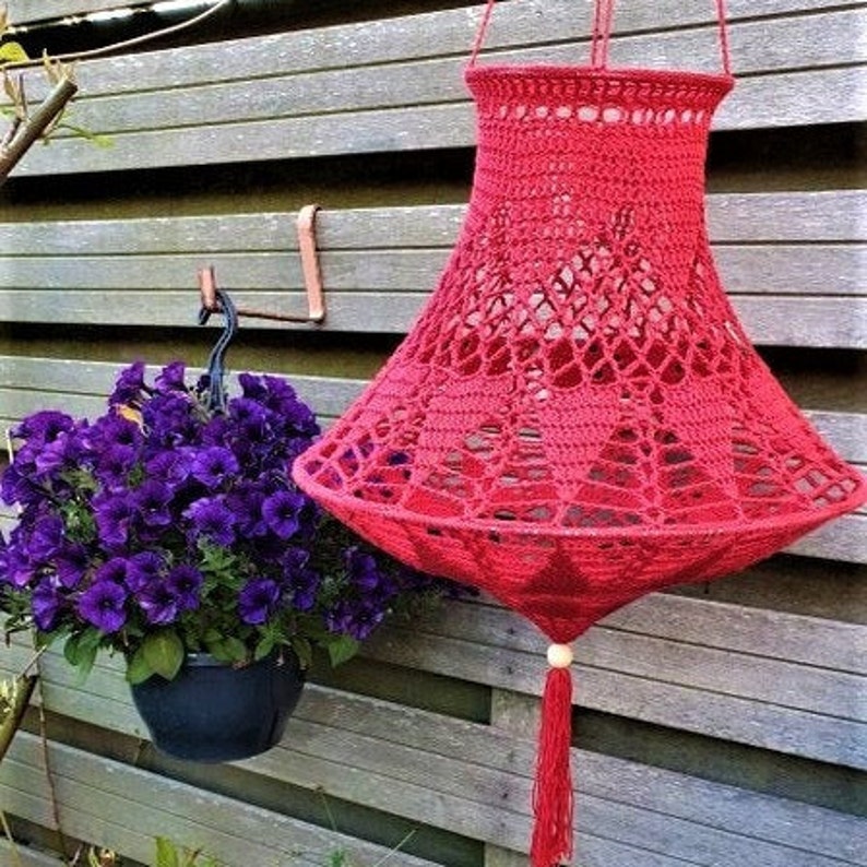 May include: A crocheted lampshade in a bright red color hangs from a hook. The lampshade has a delicate, lacy design and a tassel at the bottom.
