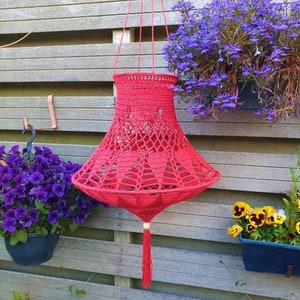 May include: A red crocheted hanging lampshade with a tassel. The lampshade is hanging from a hook on a wooden fence. There are flowers in pots on either side of the lampshade.