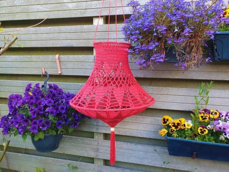 May include: A red crocheted hanging lampshade with a tassel. The lampshade is hanging from a hook on a wooden fence. There are flowers in pots on either side of the lampshade.