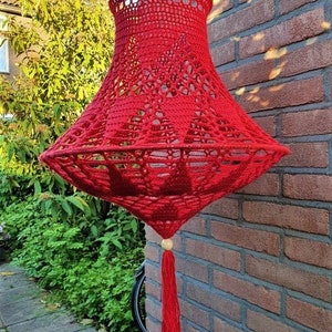 May include: A red crocheted lampshade with a tassel hanging from a wooden bead. The lampshade is hanging from a brick wall.