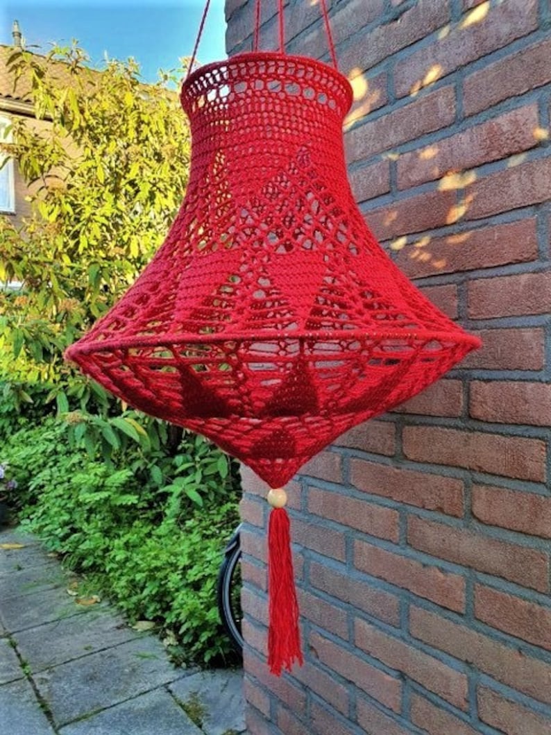 May include: A red crocheted lampshade with a tassel hanging from a wooden bead. The lampshade is hanging from a brick wall.