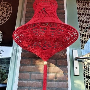 May include: A red crocheted lampshade with a white bead and tassel hanging from a brick wall.