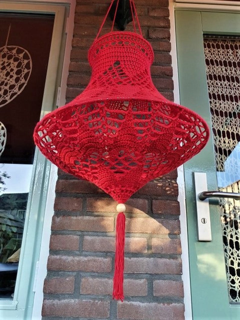 May include: A red crocheted lampshade with a white bead and tassel hanging from a brick wall.