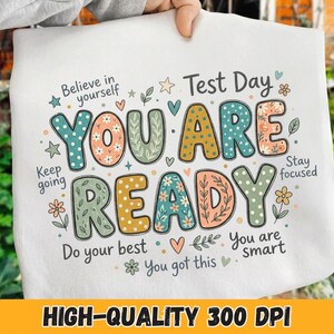Puede incluir: Camiseta gráfica blanca con las palabras "YOU ARE READY" en letras coloridas y decoradas con flores. El diseño incluye frases alentadoras como "Believe in yourself" y "You got this". También se incluyen las palabras "Test Day".