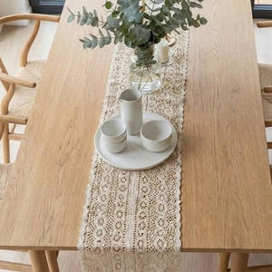 Peut inclure: Une table à manger en bois avec un chemin de table en dentelle crème. La table est ornée de bols en céramique blancs, d'une cruche et d'une assiette. Un vase en verre avec du feuillage vert et des bougies complètent le décor.