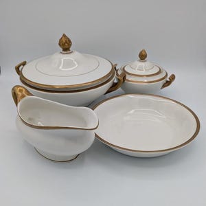 May include: A white porcelain serving set with gold trim. The set includes a lidded soup tureen, a smaller lidded dish, a gravy boat, and a serving bowl. The tureen and small dish have gold finials.