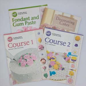 May include: A collection of Wilton cake decorating books. Titles include "Fondant and Gum Paste", "Decorating Cakes", "Course 1", and "Course 2". The covers feature images of decorated cakes and sugar flowers, with a white background.