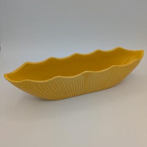 May include: A long, boat-shaped yellow ceramic bowl with a scalloped rim and a textured exterior featuring vertical, raised ridges. The vessel has a smooth, glossy finish and a deep, open interior, presenting a uniform bright yellow color throughout.