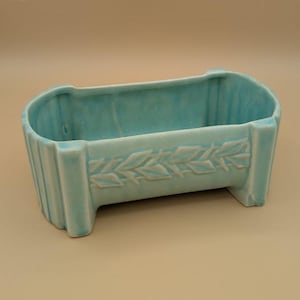 May include: A rectangular, light blue ceramic planter with a raised leaf design on the front. The planter has a ribbed texture on the sides and is supported by two rectangular feet. The overall shape is a long, shallow rectangle.