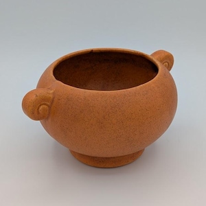 May include: A round, terracotta-colored ceramic bowl with a speckled texture, featuring two small, curled handles on opposite sides and a circular pedestal base. The interior of the bowl is smooth and deep, with a consistent matte finish throughout.