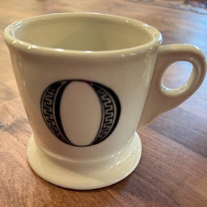 May include: Cream-colored ceramic mug with a black letter "O" in a decorative font. The mug has a handle and a wide base. The letter "O" is outlined with a Greek key design. The mug is likely for coffee or tea.