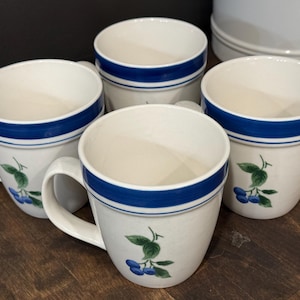 May include: A set of four off-white ceramic mugs featuring a blue band around the upper rim and a painted design of blue blueberries with green leaves on the front. Each mug has a curved handle and a smooth, rounded body shape for holding beverages.