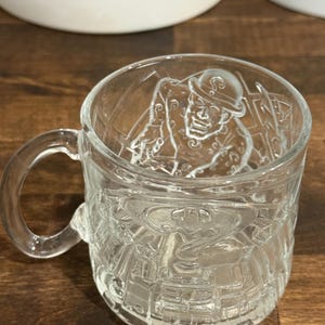 May include: A clear glass mug featuring an embossed design of a character wearing a bowler hat and a suit covered in question marks. The glass surface displays a raised relief pattern of the character and a large question mark symbol on the front.