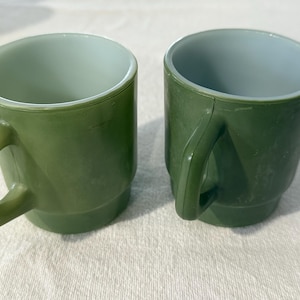 Puede incluir: Dos tazas verdes vintage con asa cuadrada e interior azul claro. Las tazas están hechas de un material duradero y están apiladas sobre una superficie blanca. Las tazas son de diseño clásico, perfectas para café o té.