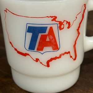 May include: A white milk glass mug featuring a red outline of the United States map. Centered on the map is a blue and red logo with the letters TA inside a shield shape. The mug has a cylindrical body with a stepped base and a side handle.