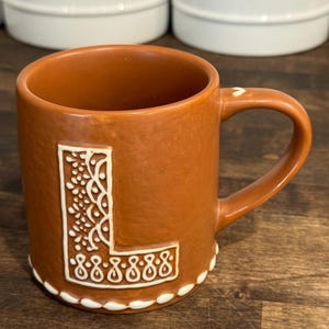 May include: A brown ceramic mug featuring a large, embossed letter L on the front. The letter is decorated with white, intricate, swirling patterns and dots. A white, scalloped, icing-like trim runs along the base of the mug and on the handle.