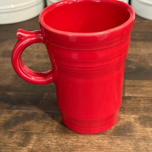 May include: A bright red ceramic mug with a tall, cylindrical body and a curved handle. The exterior features a series of horizontal embossed rings around the upper and lower sections, providing a textured appearance to the smooth, glossy finish.