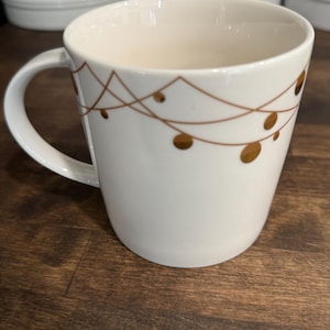 May include: A white ceramic mug featuring a delicate, thin brown line pattern that mimics string lights draped around the upper portion of the vessel, accented with small, solid brown circular dots representing light bulbs.