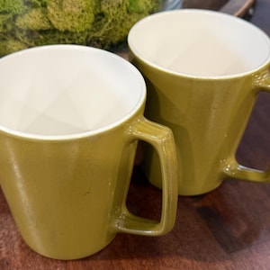 May include: Two olive green mugs with angular handles and off-white interiors are displayed. The mugs are made of a solid material and have a geometric design. The mugs are on a dark wooden surface.