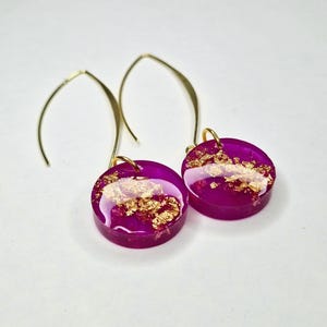 May include: A pair of magenta resin earrings with gold-colored accents. The earrings have a circular design with gold leaf embedded in the resin. The ear wires are gold-colored and curved.