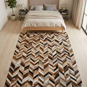 Op de afbeelding: Een rechthoekig vloerkleed met een visgraatpatroon bestaande uit individuele stroken koeienhuid in tinten bruin, zwart, wit en beige. Het kleed ligt op een lichte houten vloer voor een bed met een beige gestoffeerd hoofdeinde.