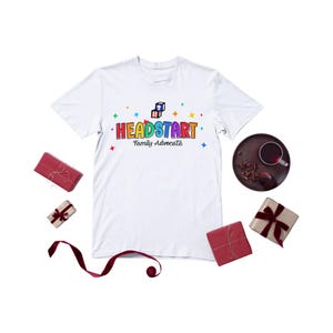 Puede incluir: Camiseta blanca con el texto "HEADSTART Family Advocate" en letras coloridas y estrellas. La imagen incluye regalos envueltos en rojo y marrón, un plato oscuro con una taza de té y una cinta granate.
