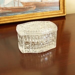 May include: A clear, oval-shaped glass trinket box with a removable lid. The exterior features a pressed geometric pattern of small squares and faceted textures that catch the light. The lid has a matching textured design with a smooth, flat top.