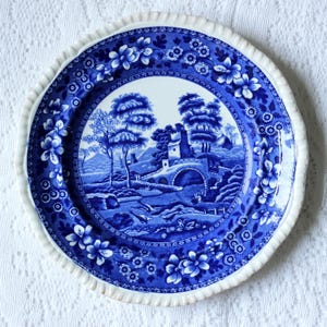 May include: A round ceramic plate featuring a blue and white transferware pattern. The center depicts a landscape with a stone bridge, trees, and buildings. The wide rim is decorated with a dense floral motif and has a scalloped, fluted edge.