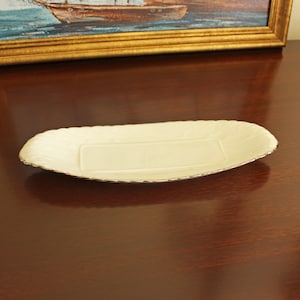 May include: An oval-shaped cream-colored ceramic serving dish featuring a smooth central rectangular panel and a textured, scalloped rim accented with a thin gold-toned metallic trim along the outer edge of the piece.
