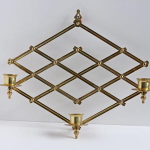 Könnte beinhalten: Ein Vintage-Wandleuchter aus Messing mit rautenförmigem Design. Der Leuchter verfügt über drei Kerzenhalter und ein dekoratives, geometrisches Muster. Das Messing hat einen warmen, goldenen Farbton und verleiht dem Stück einen Hauch von Eleganz.