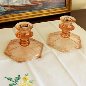 May include: Two vintage, peach-colored glass candle holders. Each holder has a hexagonal base and a detailed, tiered stem leading to a circular top. The candle holders are displayed on a white cloth with embroidered flowers, with a framed painting in the background.