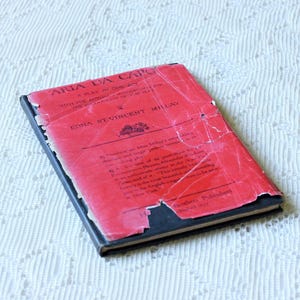 May include: A vintage book titled Aria Da Capo by Edna St. Vincent Millay with a distressed red paper cover featuring black text and a small floral illustration. The book has a black spine and edges, showing significant wear and creasing.
