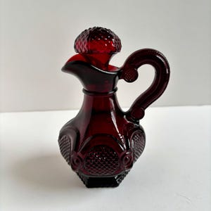 May include: A ruby red glass cruet with a handle and a stopper. The cruet has a textured pattern on the body and a decorative handle. The stopper has a textured, rounded top. The cruet is likely used for serving oil or vinegar.