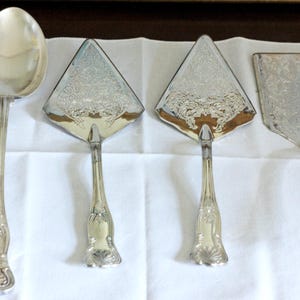 May include: A set of four silver-toned serving utensils including one large oval spoon and three flat serving pieces with triangular and rectangular heads. Each handle features an ornate embossed pattern with a shell motif at the base.