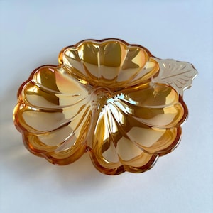 May include: An iridescent amber-colored glass dish in the shape of a leaf. The dish has a scalloped edge and a detailed leaf design on the side. The glass reflects light, creating a shimmering effect.