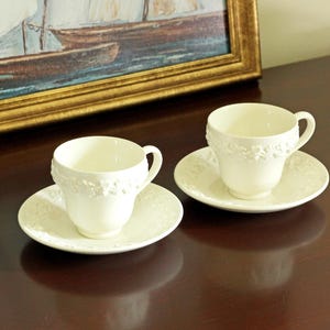 May include: Two cream-colored porcelain teacups and matching saucers featuring a raised, embossed floral relief pattern around the rims. Each cup has a curved handle and a smooth interior, while the saucers display a coordinating decorative border.