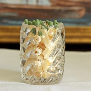 May include: A clear glass match holder with a textured, spiral-ribbed exterior and a scalloped top edge. The cylindrical vessel is filled with wooden matches that have light green tips, visible through the transparent, patterned glass walls.