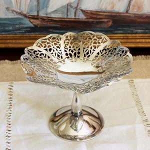 May include: A silver-colored, ornate pedestal bowl with a scalloped edge and intricate openwork design. The bowl sits on a short stem and a round base. The base has a leaf detail. The background features a painting of sailboats.