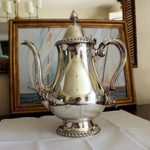 May include: A polished silver coffee pot with a decorative handle and spout. The pot has a lid with a small finial and sits on a round base. The pot reflects the surrounding room and is placed on a white cloth.