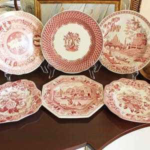May include: A collection of six ceramic plates featuring red and white transferware patterns. The set includes three round plates with detailed landscape and floral motifs, one octagonal plate with a river scene, and two plates with scalloped edges.