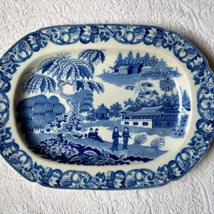 May include: An oval ceramic platter featuring a blue and white transferware pattern. The central scene depicts figures, trees, and buildings in a landscape, while the wide border is decorated with an intricate floral and scrollwork design.