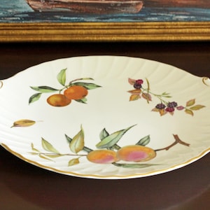 May include: A white ceramic serving platter with a scalloped edge and gold-trimmed handles. The surface features painted illustrations of oranges, blackberries, and peaches with green leaves. The platter has a ribbed texture along the rim.