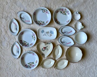 May include: A collection of various ceramic dishes including round bowls with floral patterns, oval plates with geometric accents, small scalloped dishes, a heart-shaped plate with a bird illustration, and a rectangular plate with blue flowers.