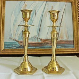May include: A pair of polished brass candlesticks featuring a tall, slender stem with a flared top cup and a wide, hexagonal base. The metallic surface reflects light, highlighting the smooth, tapered design of these two matching candle holders.