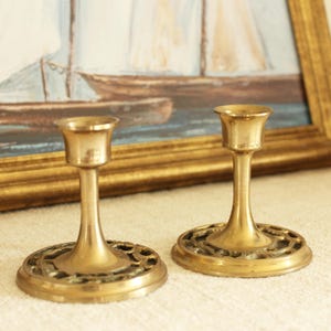 Könnte beinhalten: Zwei antike Kerzenhalter aus Messing. Jeder Halter hat einen runden Sockel mit einem verzierten Design, einen schlanken Stiel und eine ausgestellte Oberseite für eine Kerze. Die Kerzenhalter werden vor einem gerahmten Gemälde präsentiert.